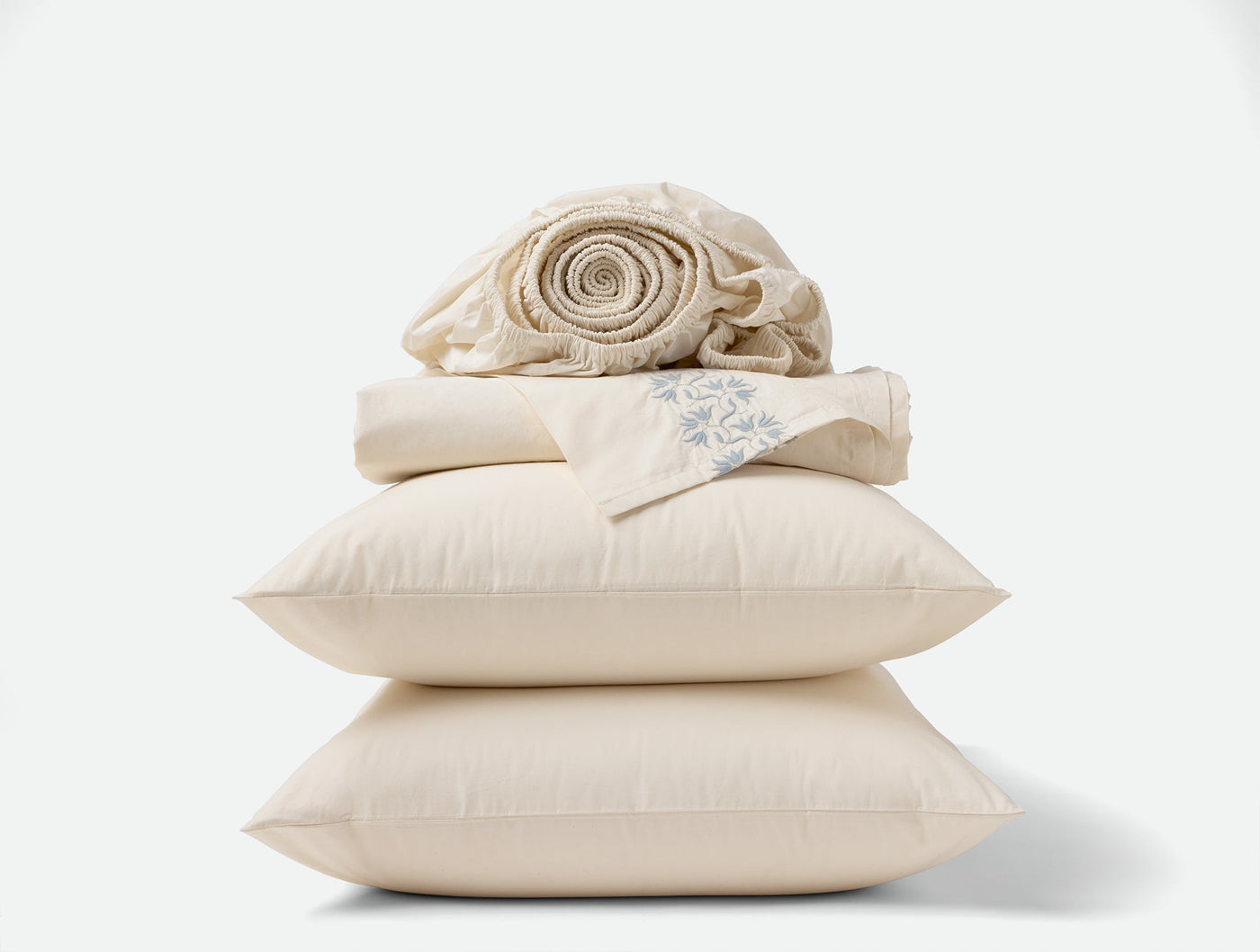 Close-up of stacked ivory Coyuchi Refined Percale pillows and folded sheets on white background.