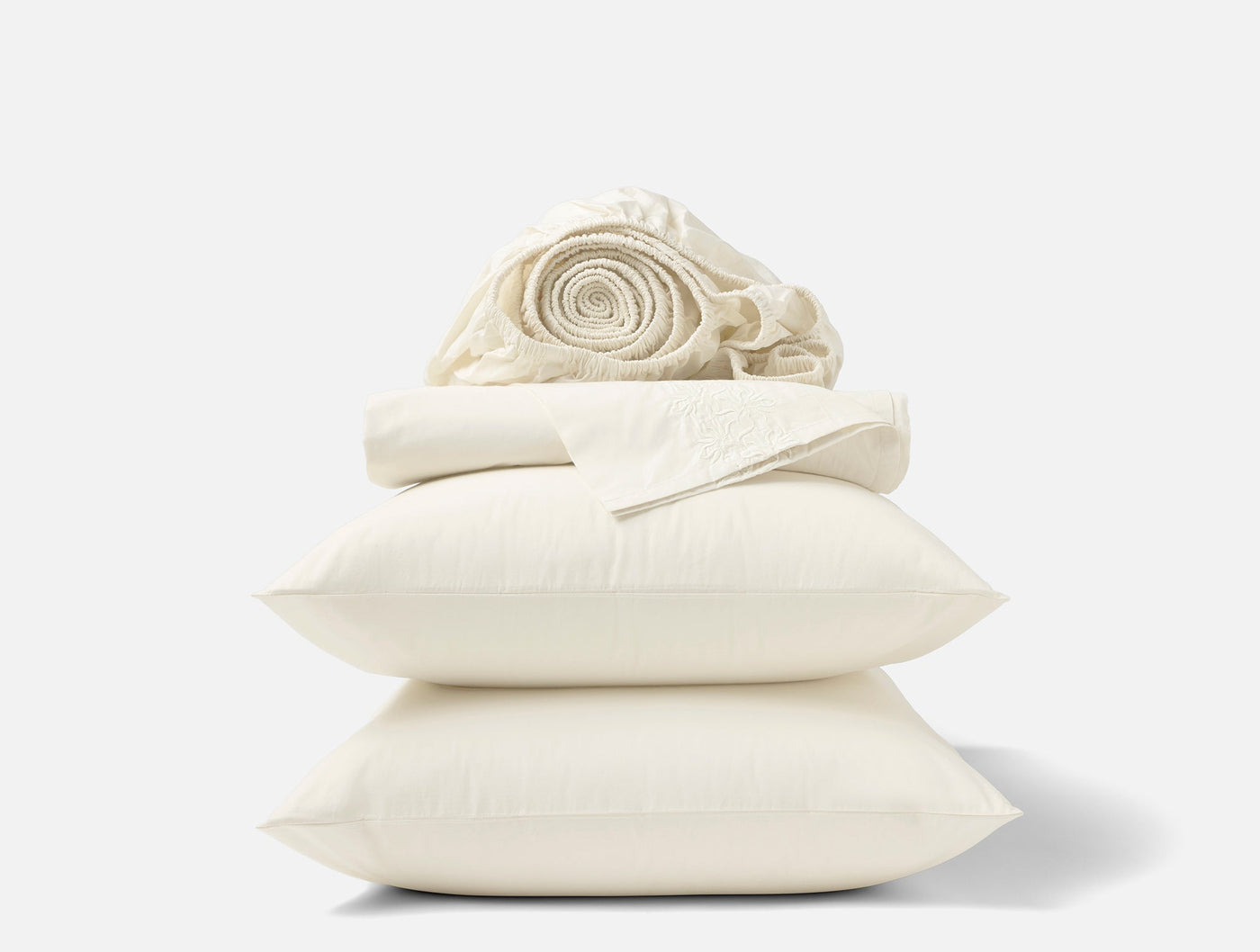 Coyuchi Refined Percale sheet set styled as stacked ivory pillows with rolled flat sheet on top.