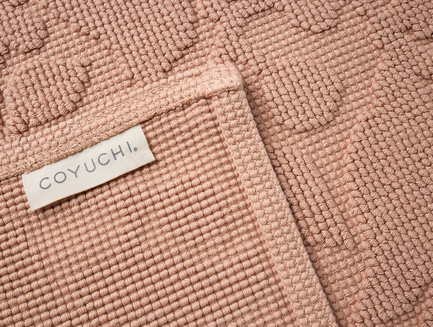 Close-up of Coyuchi Wildflower bath rug corner in blush, showing tufted texture and label.