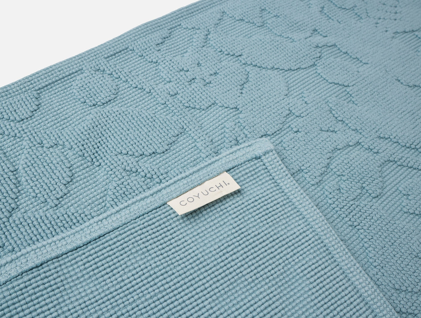 Close-up of Coyuchi Wildflower bath rug corner in slate blue, showing floral texture and woven label.