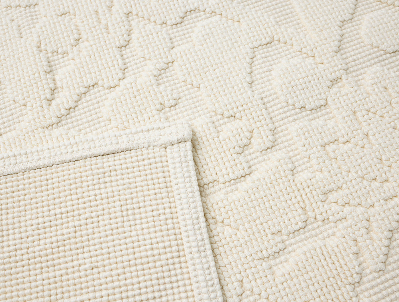Close-up of ivory Coyuchi Wildflower bath rug corner, showing textured floral pattern and folded edge.