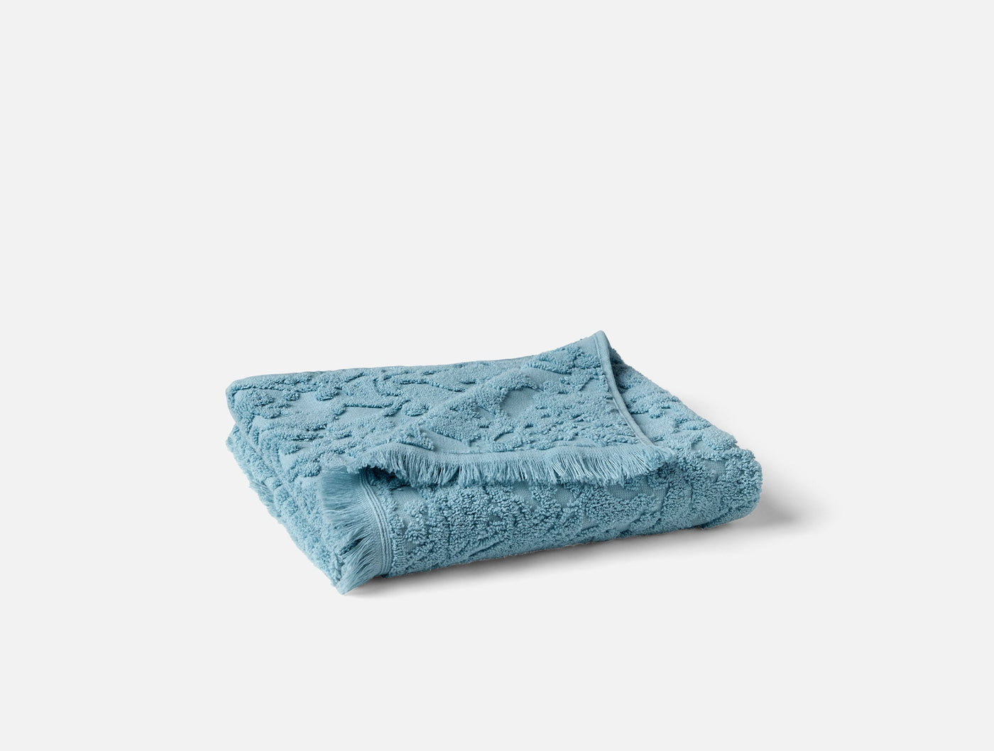 Folded cerulean bath towel showing sculpted wildflower jacquard texture and fringe hem detail.
