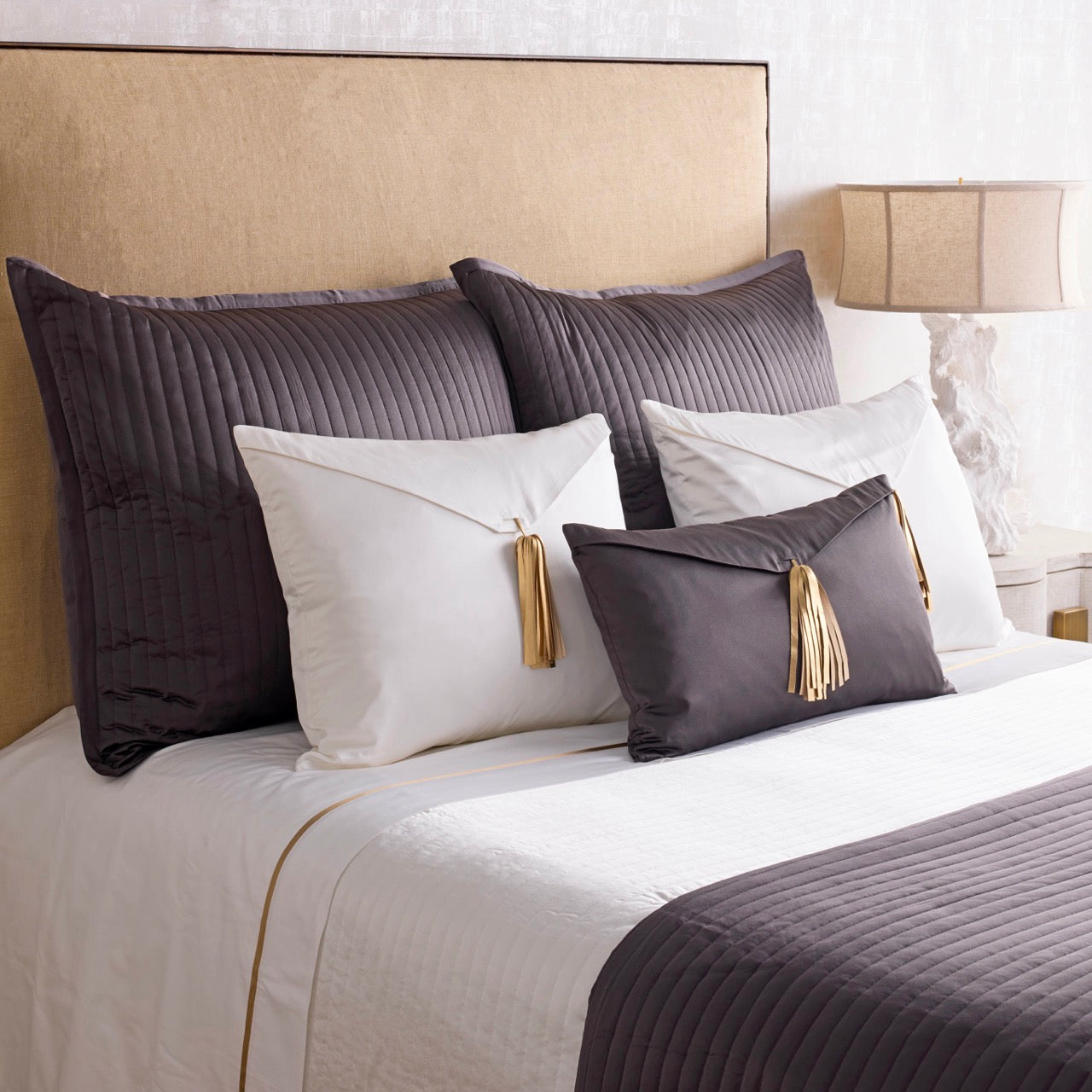 Charcoal velvet Saihai pillow with gold tassel, styled on a layered bed with ivory bedding.
