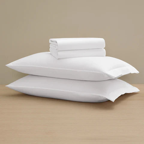 Two Frette Sateen | Pillowcase Sets by Frette are stacked on a wooden surface with a folded sheet set on top, all against a beige background—a luxurious bedding upgrade.