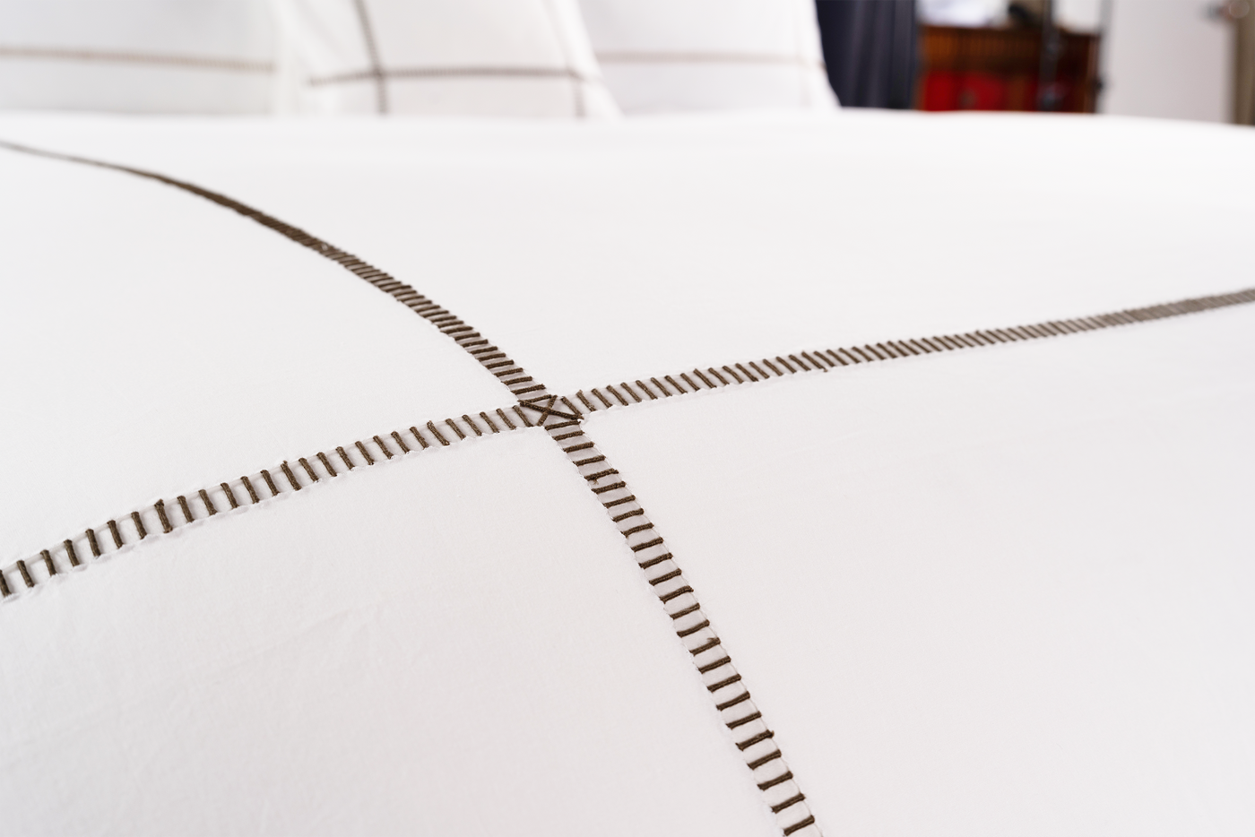 Close-up of intersecting dashed embroidery in taupe on white cotton bedding.