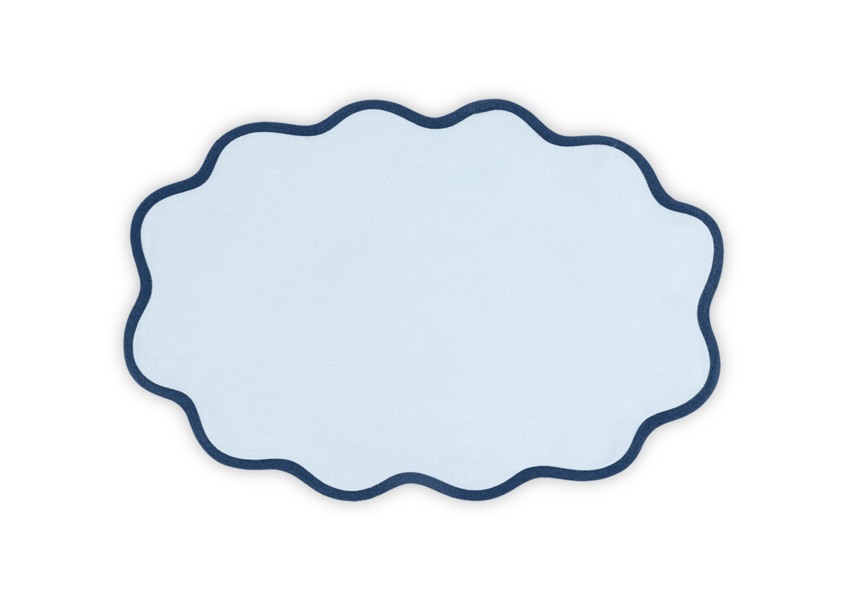 The Matouk Scallop Edge Placemat Set features light blue linen with a scalloped edge and dark blue border, bringing charm to any tablescape when set against a white background.
