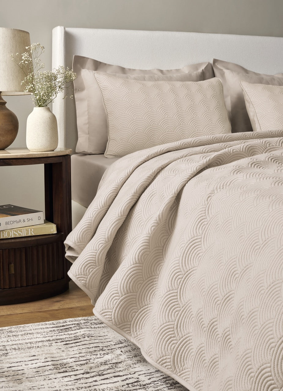Ann Gish Scallop coverlet set in sand, styled on bed with arc quilting detail and matching shams.