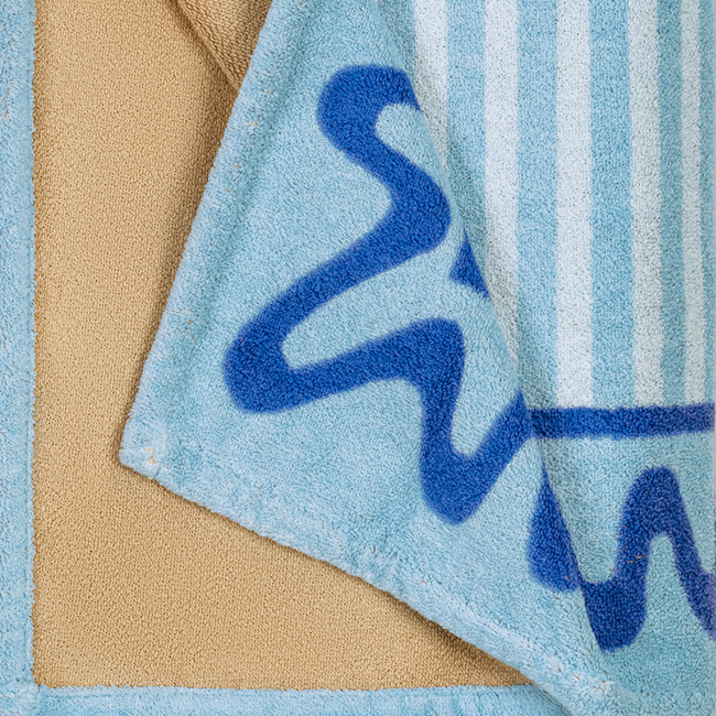A close-up of the Matouk Seahorse Beach Towel, featuring a wavy dark blue pattern and vertical stripes on soft cotton terry, partially folded atop a beige textured surface.