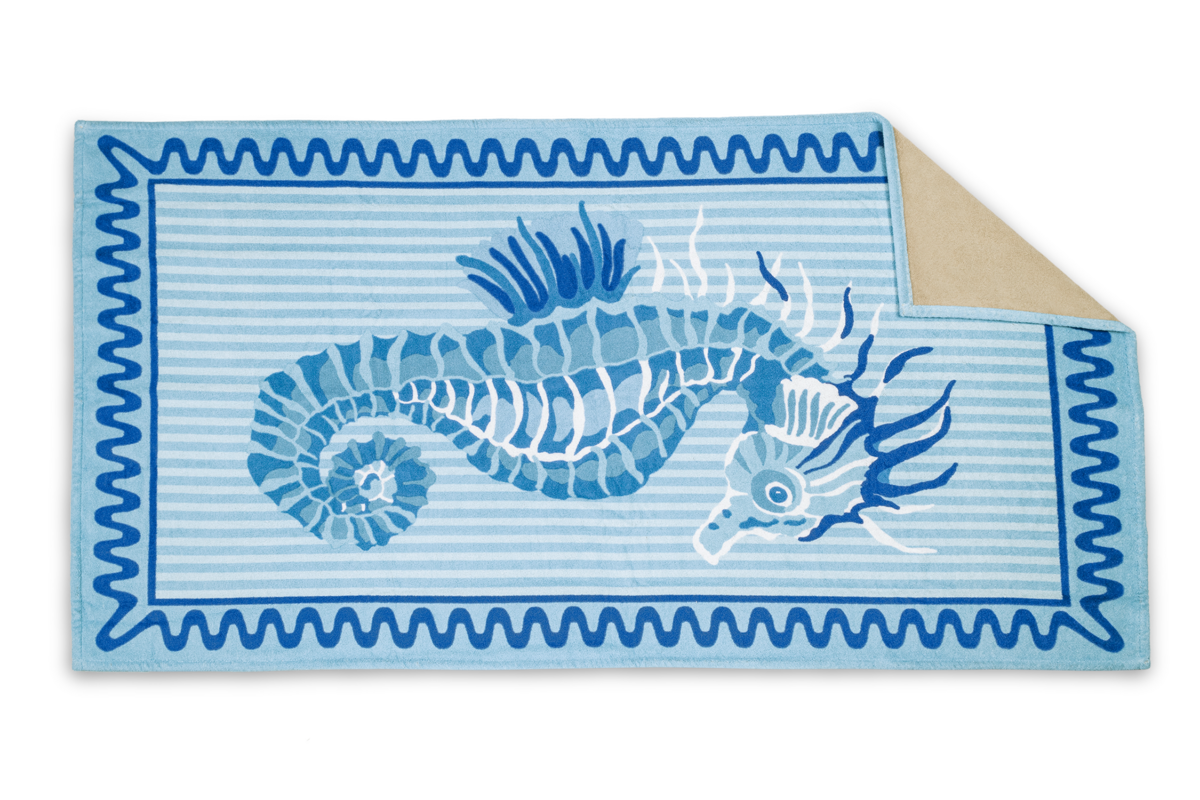 The Matouk Seahorse Beach Towel is a rectangular blue and white striped towel with a central seahorse illustration and a solid beige folded corner.