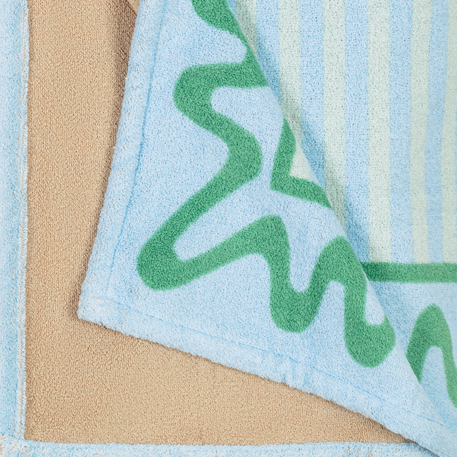 A close-up of the Seahorse Beach Towel by Matouk shows its blue and green pattern with a beige cotton terry underside—a stylish piece from the Schumacher collaboration.