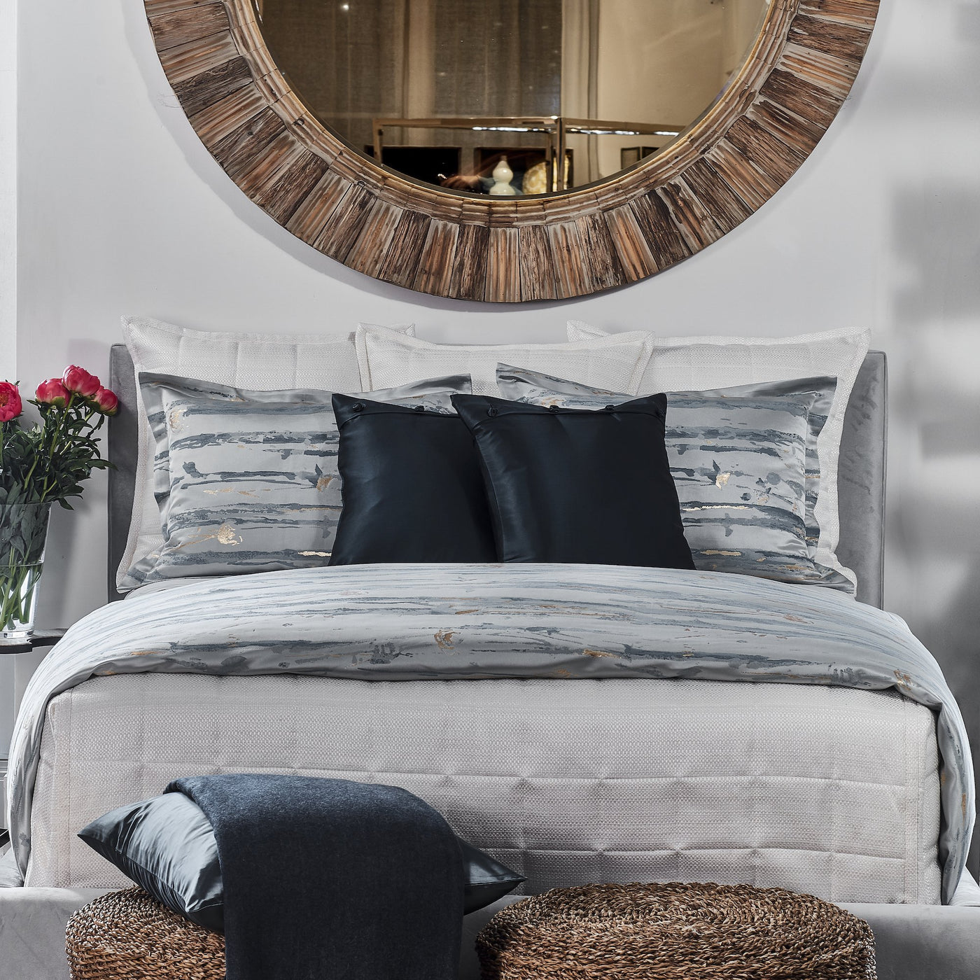 Styled bed with Ann Gish Serenity duvet, layered grey and black pillows, round wood mirror above.
