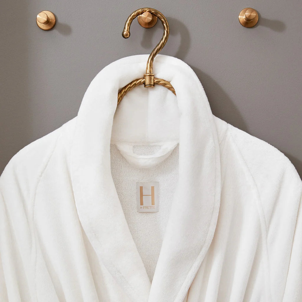 A Frette Shawl Collar Robe hangs on a gold hook against a gray wall, with an H label stitched on the inside collar.
