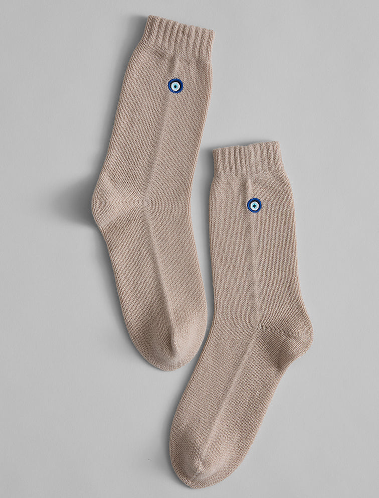 The Matouk Sleeping Socks by Matouk are beige cozy socks featuring a small blue and white circular emblem near the top, shown laid flat on a light gray background.
