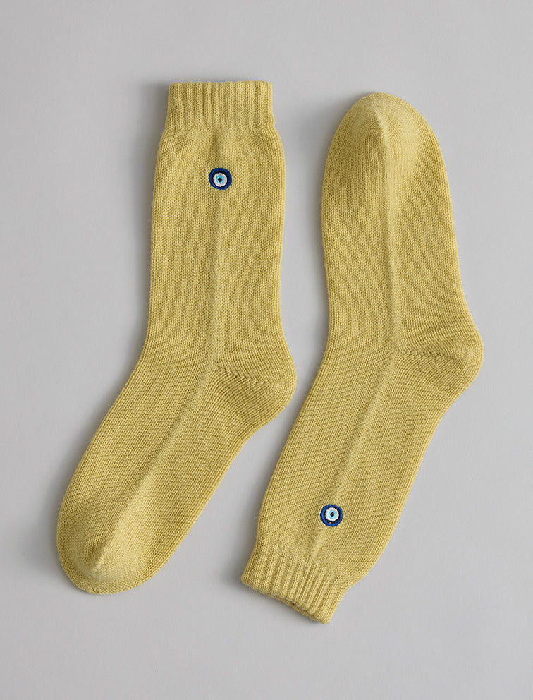 The Matouk Sleeping Socks by Matouk are cozy yellow socks with ribbed cuffs and a small blue and white circular emblem near the ankle, made in Scotland and shown on a light grey background.