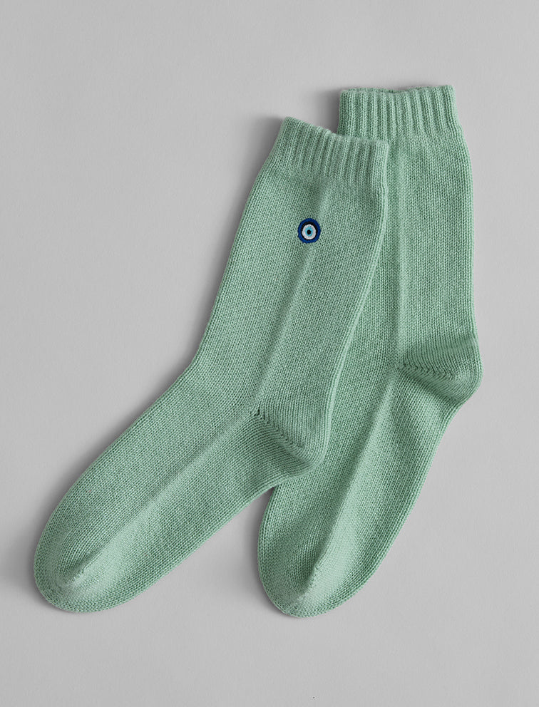The Matouk Sleeping Socks by Matouk are light green cozy socks with ribbed cuffs and a blue-white circular emblem, perfect for lounging or wearing as cashmere sleeping socks.