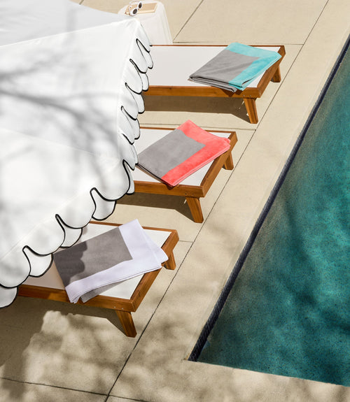 Three Peacock Alley Soleil beach towels in colorblock designs draped on teak pool loungers poolside.
