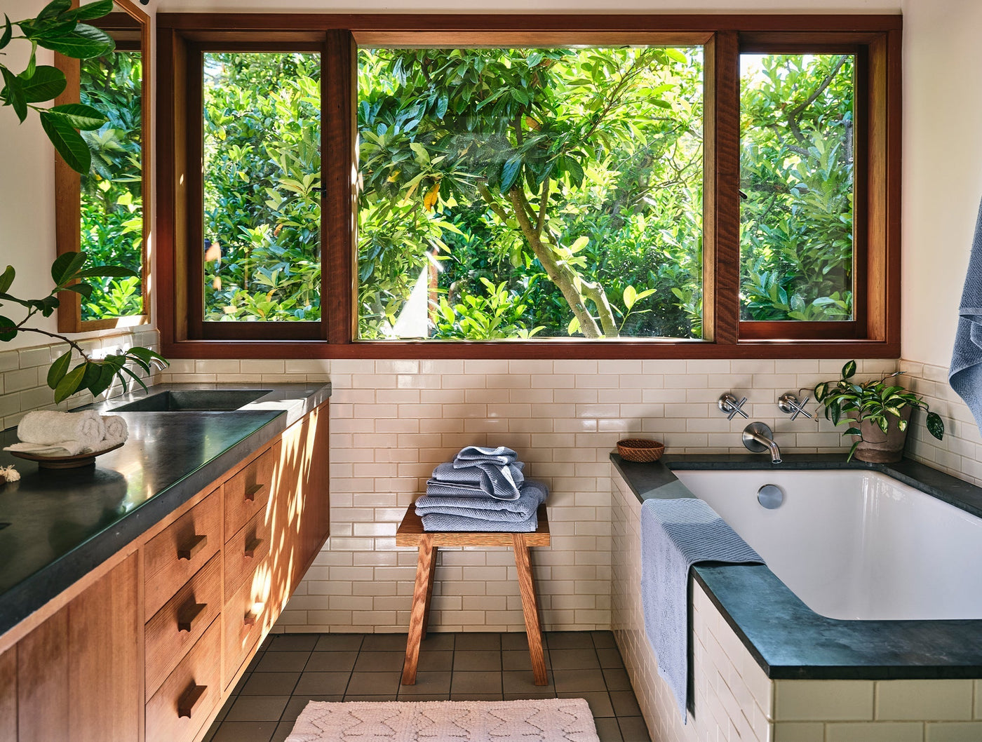 Folded slate blue Coyuchi Air Weight bath sheets stacked on a wooden stool in a sunlit bathroom.