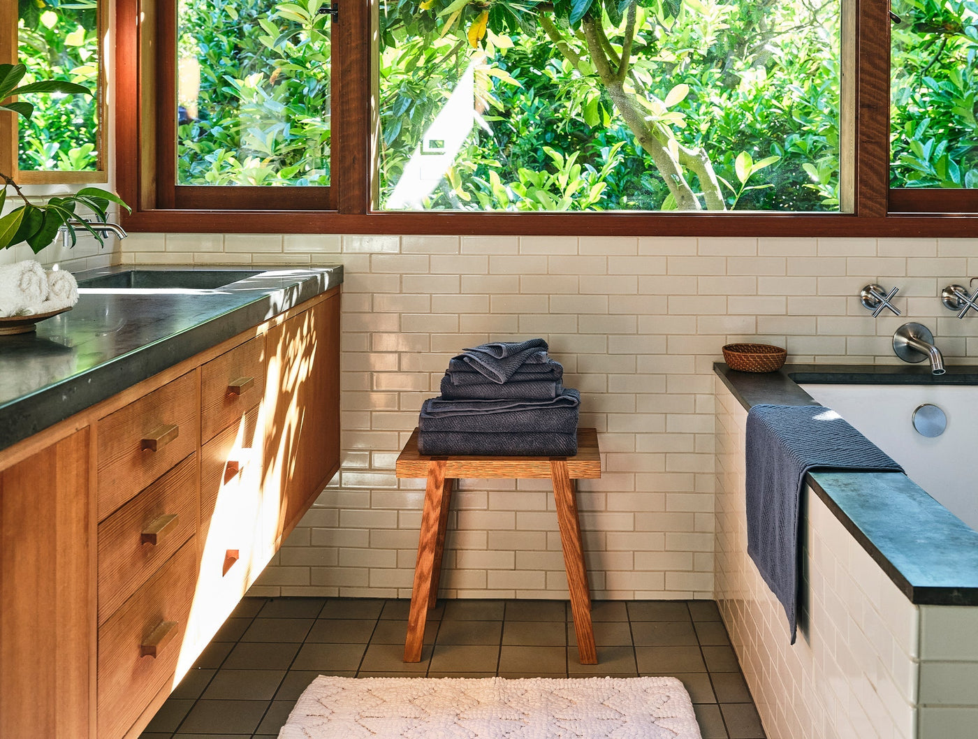 Folded slate blue Coyuchi Air Weight bath towels stacked on a wooden stool in a sunlit bathroom.