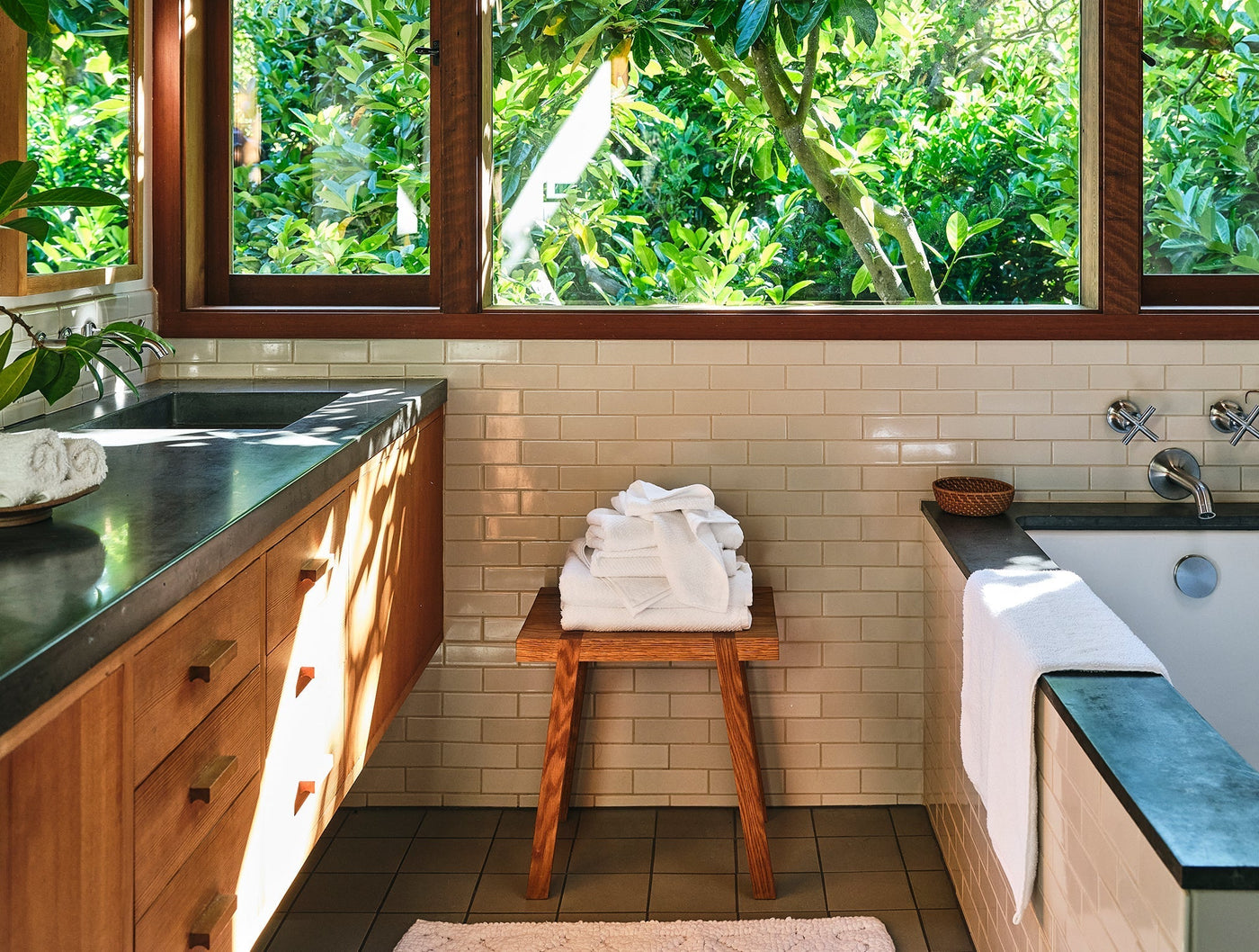 Folded white Coyuchi Air Weight wash cloths stacked on a wooden stool in a sunlit bathroom.
