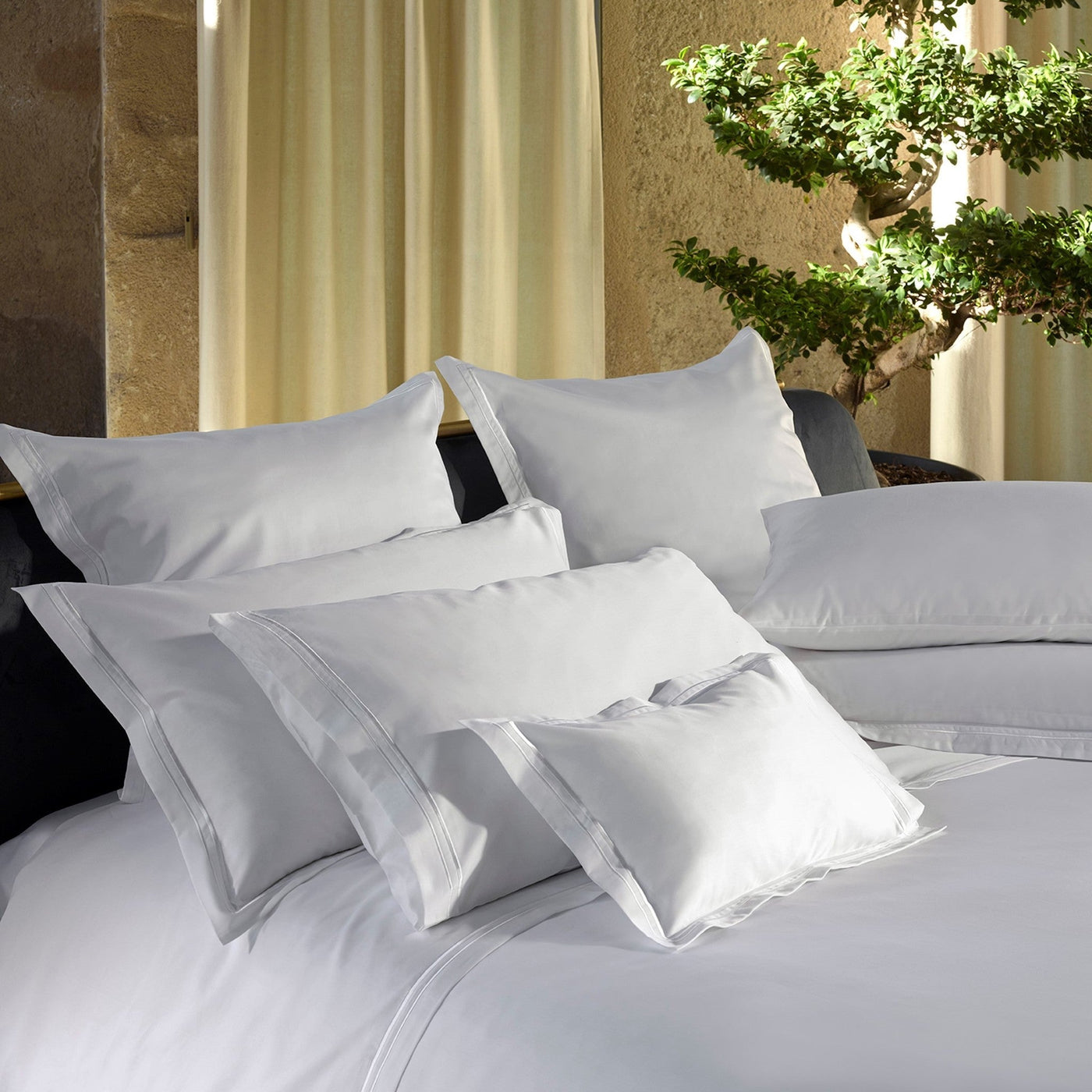 White Sublime pillow shams styled on a made bed with layered bedding and a bonsai tree backdrop.