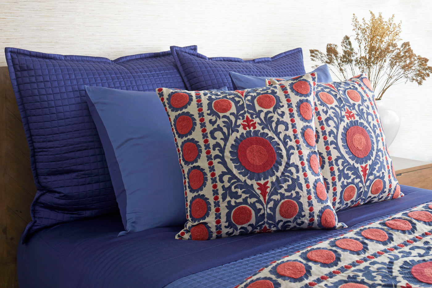 Embroidered suzani pillow with red floral medallions on ivory and navy, styled on a blue bed.