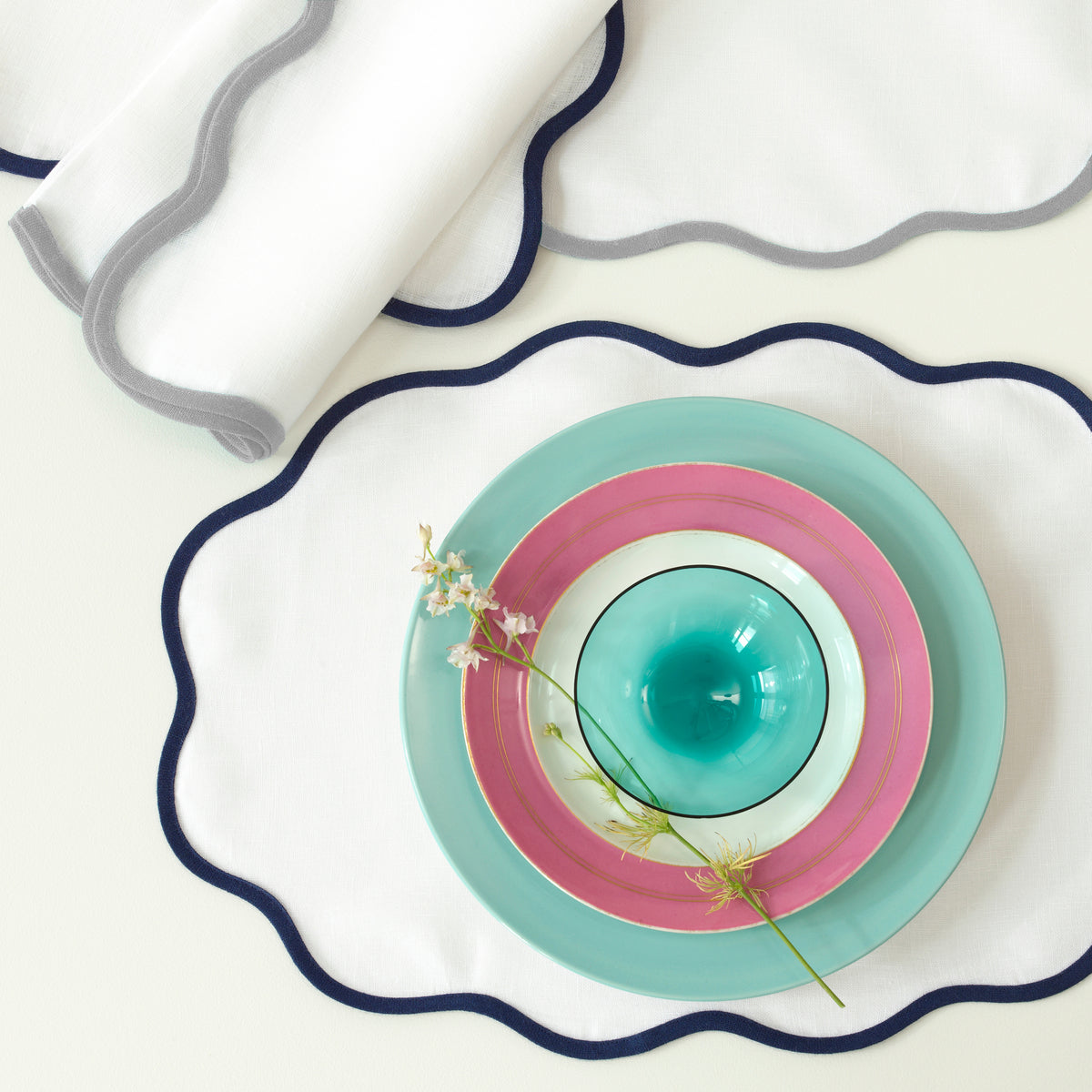 A unique tablescape features Matouk's Scallop Edge | Napkin Set with linen placemats, pastel turquoise and pink plates and bowl, topped with a small sprig of flowers.