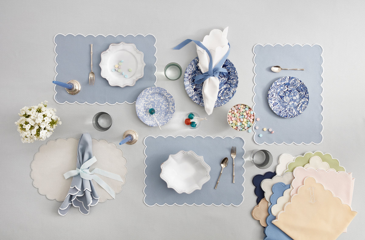 A neatly arranged table featuring Matouk Savannah Gardens placemats, assorted blue and white plates, cutlery, glasses, a small floral bouquet, candy, and folded ribbon-tied napkins.