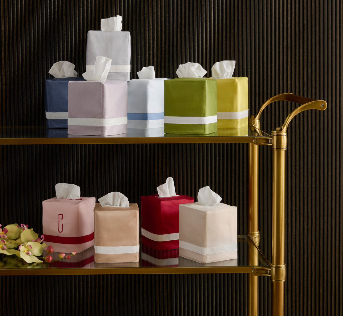A selection of colorful, square and rectangular Matouk tissue box covers—including the Nocturne | Tissue Box Cover—are arranged on a gold and glass shelf, bringing a stylish accent to a dark, vertically-lined backdrop.