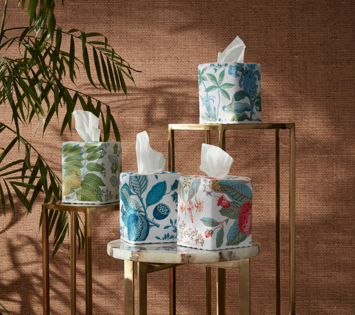 Four Matouk Pomegranate | Linen Tissue Box Covers with floral designs are displayed on marble and metal stands, with a plant and a textured brown wall in the background.