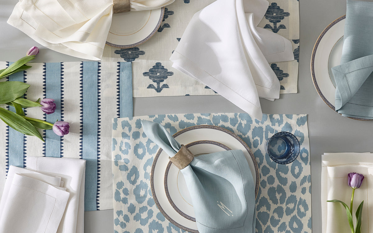A table set with a Matouk Valentina Hemstitch tablecloth, blue-patterned placemats, white and light blue napkins, gold-rimmed plates, clear glasses, and purple tulips on a gray surface.