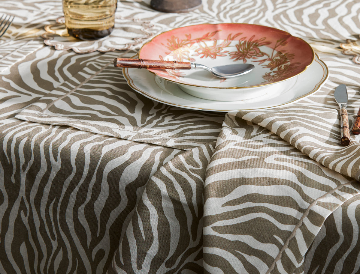 A close-up of a table set with a decorative plate, soup spoon, and glass atop Matouk's Regine Placemat Set, featuring a stylized zebra motif.
