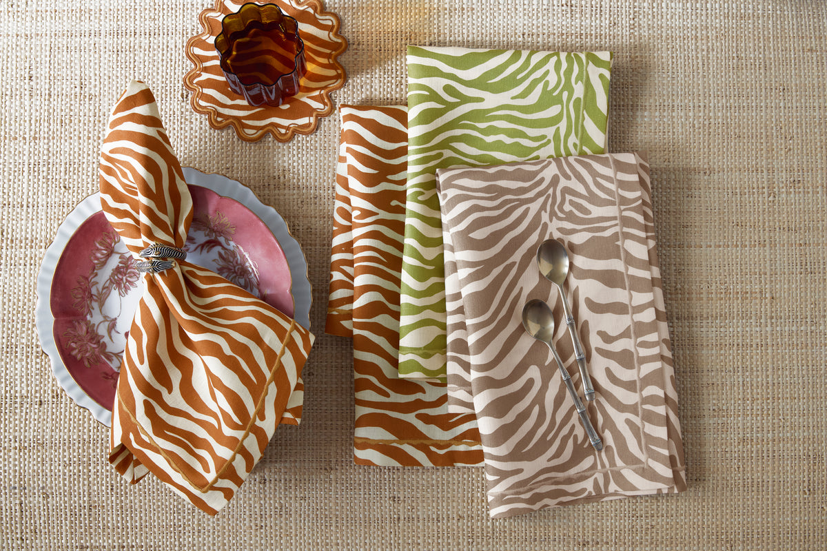 The Regine | Napkin Set by Matouk features decorative linen napkins in brown, green, and beige zebra print, shown with a plate, napkin ring, glass, and spoons on a woven placemat.