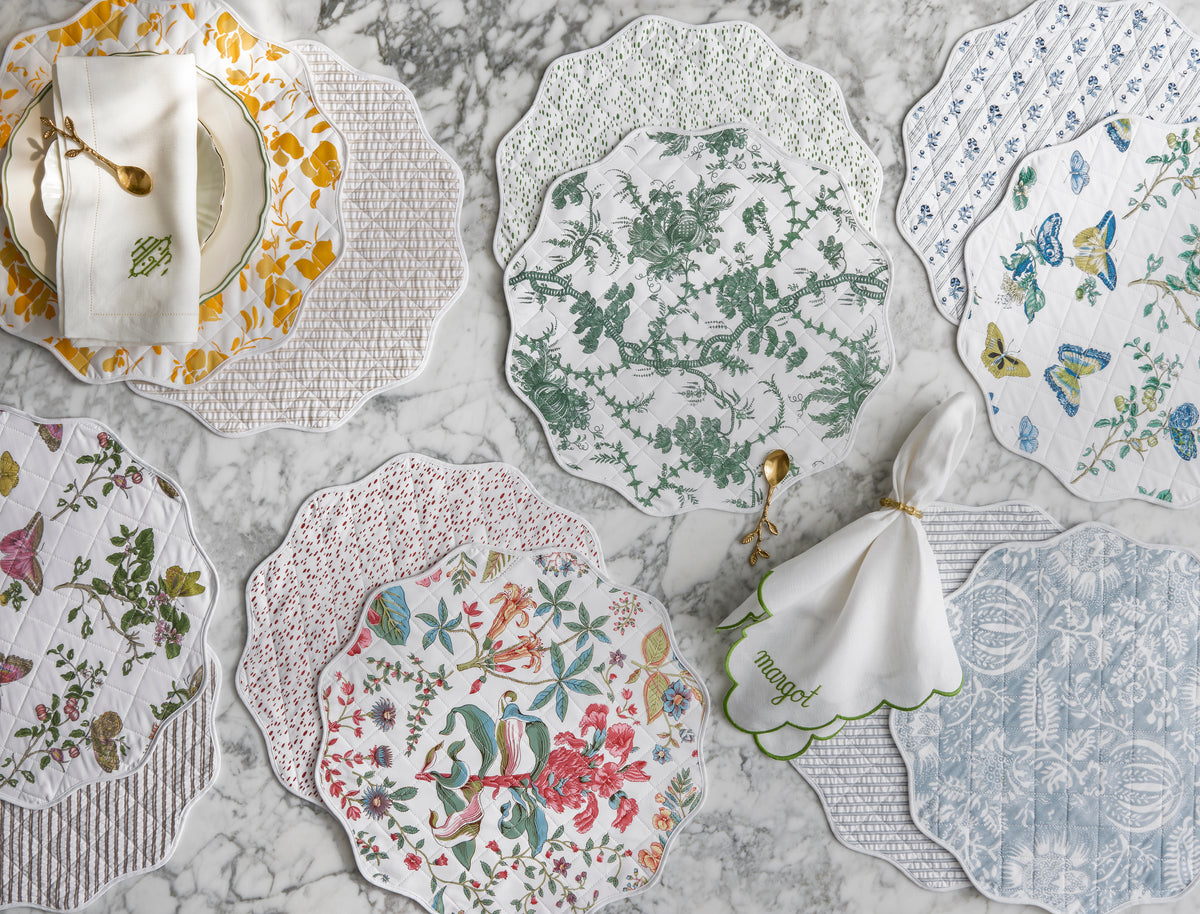 The Matouk Quilted Reversible Placemat Set features eight scalloped placemats with colorful floral and botanical designs, arranged on marble for a vibrant dining table look with plates, napkins, and gold-toned flatware.