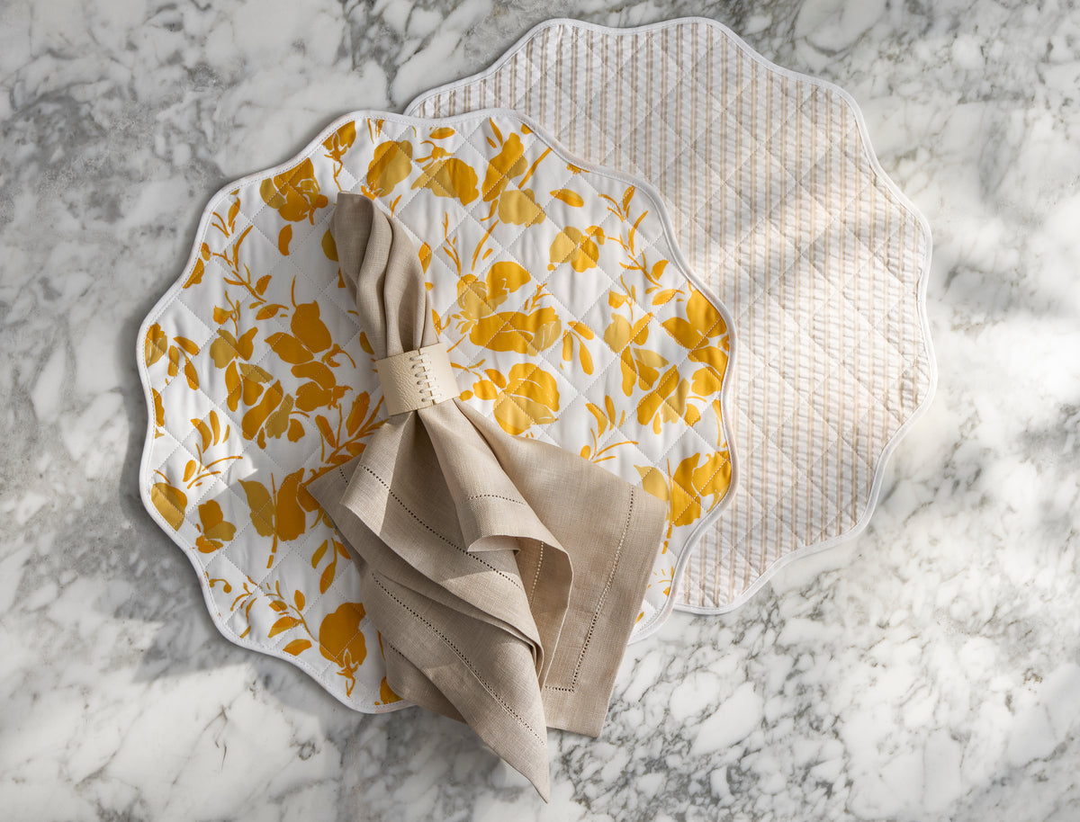 Two Quilted Reversible Placemats by Matouk rest on marble—one with a yellow floral pattern, the other beige and white. A beige napkin sits atop the floral mat, adding elegance and charm to your dining table decor.