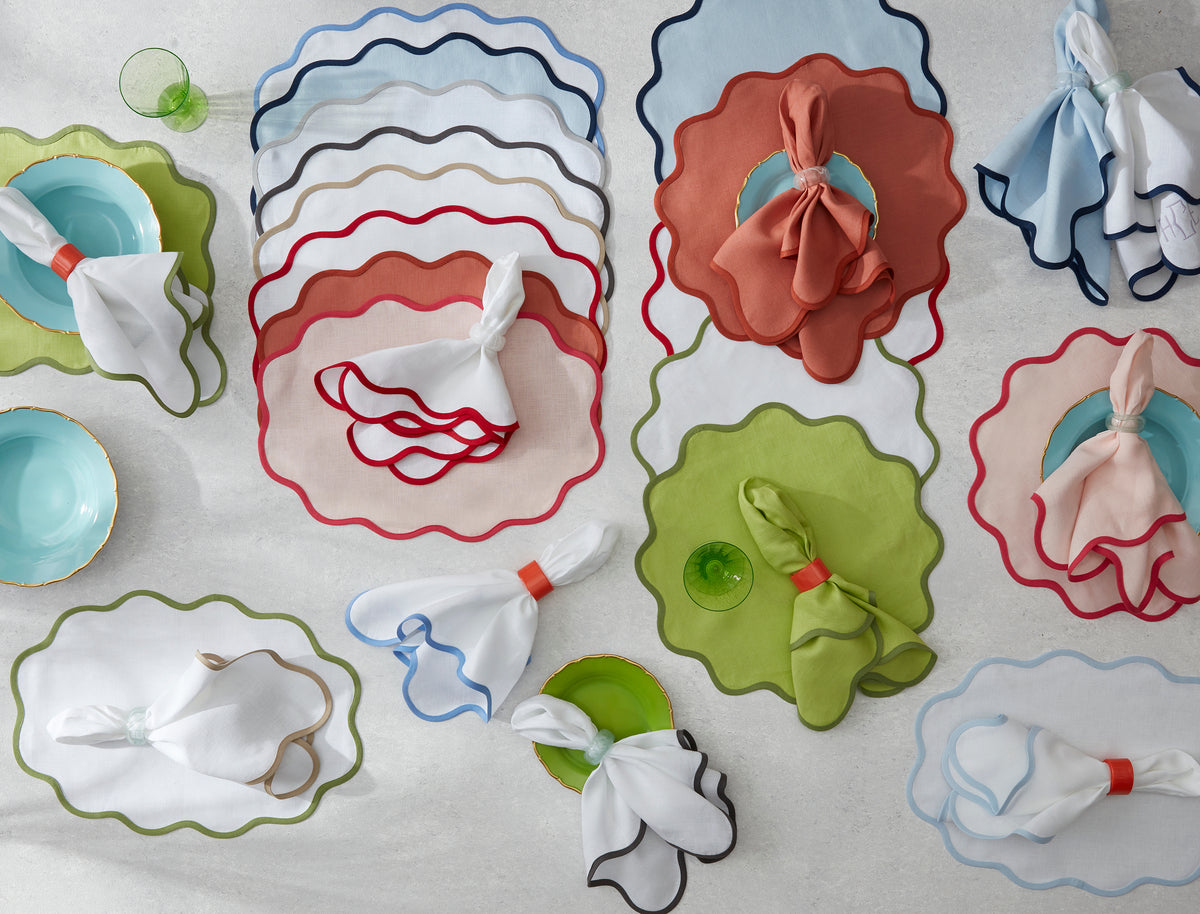 Matouk's Scallop Edge | Napkin Set in assorted colors is arranged with linen placemats, blue plates, and green glasses on a tabletop for a distinctive tablescape.