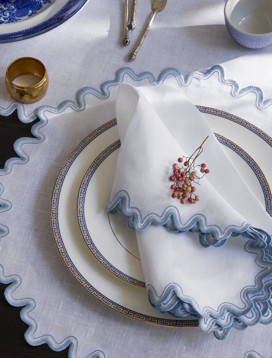 A Matouk Wave linen guest towel with blue scalloped trim and dried berries sits on a white Greek key plate atop a matching placemat. Silverware and a gold napkin ring complete this elegant, embroidered tabletop setting.