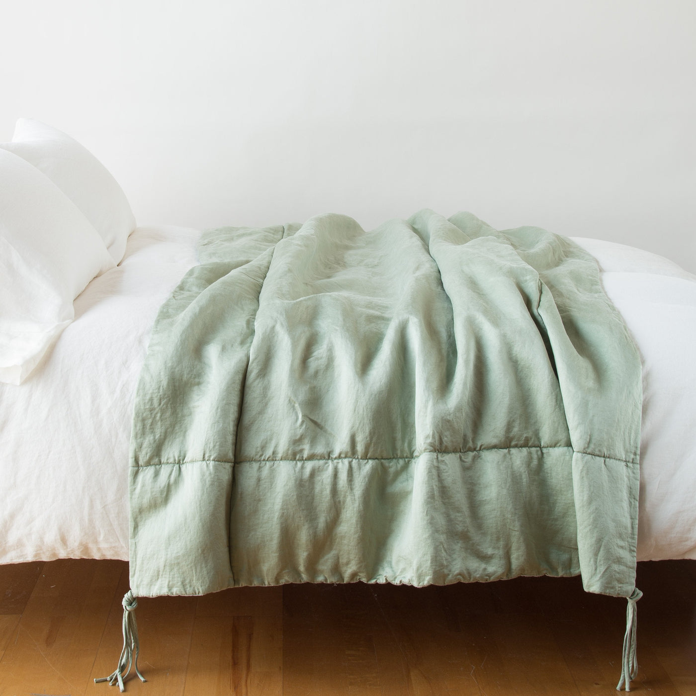 Eucalyptus linen throw blanket draped across a white bed, tassel corner detail visible.