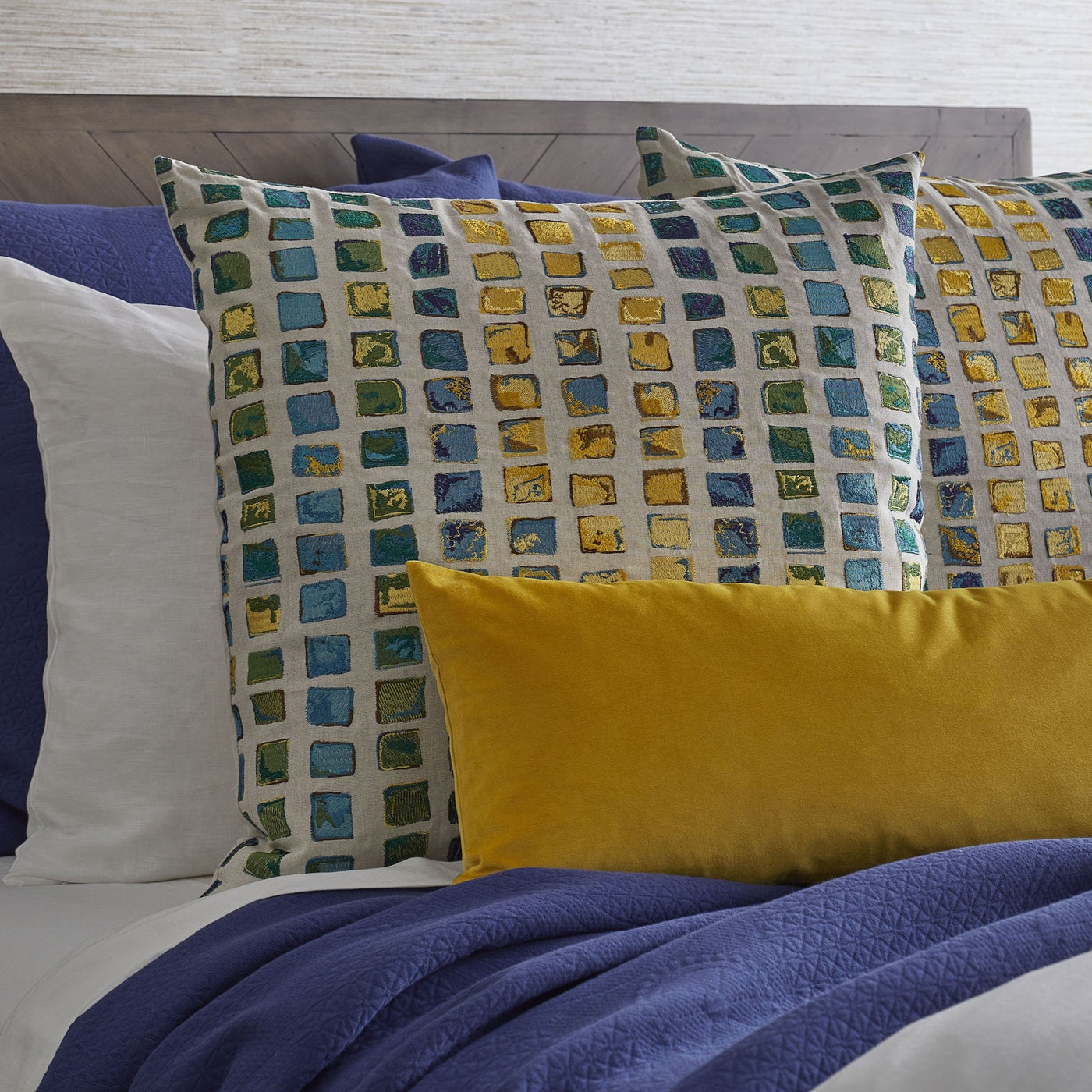 Ann Gish Tesserae Mosaic pillow with teal, green, and gold tile pattern, styled on blue bedding.