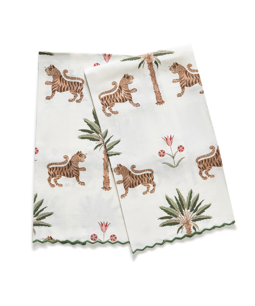 The Matouk Tiger Palm Guest Towel Set features white towels with Schumacher’s printed tigers, palm trees, and Ottoman florals, detailed with scalloped edges and green trim.