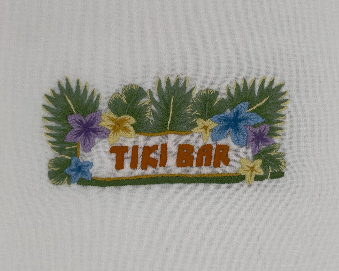 Close-up of embroidered Tiki Bar motif with tropical flowers and palm leaves on white linen.