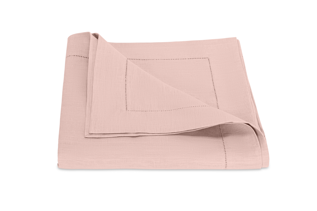 The Matouk Valentina Hemstitch Tablecloth in light pink linen, featuring a decorative hemstitch border, is neatly folded and displayed on a plain white background.
