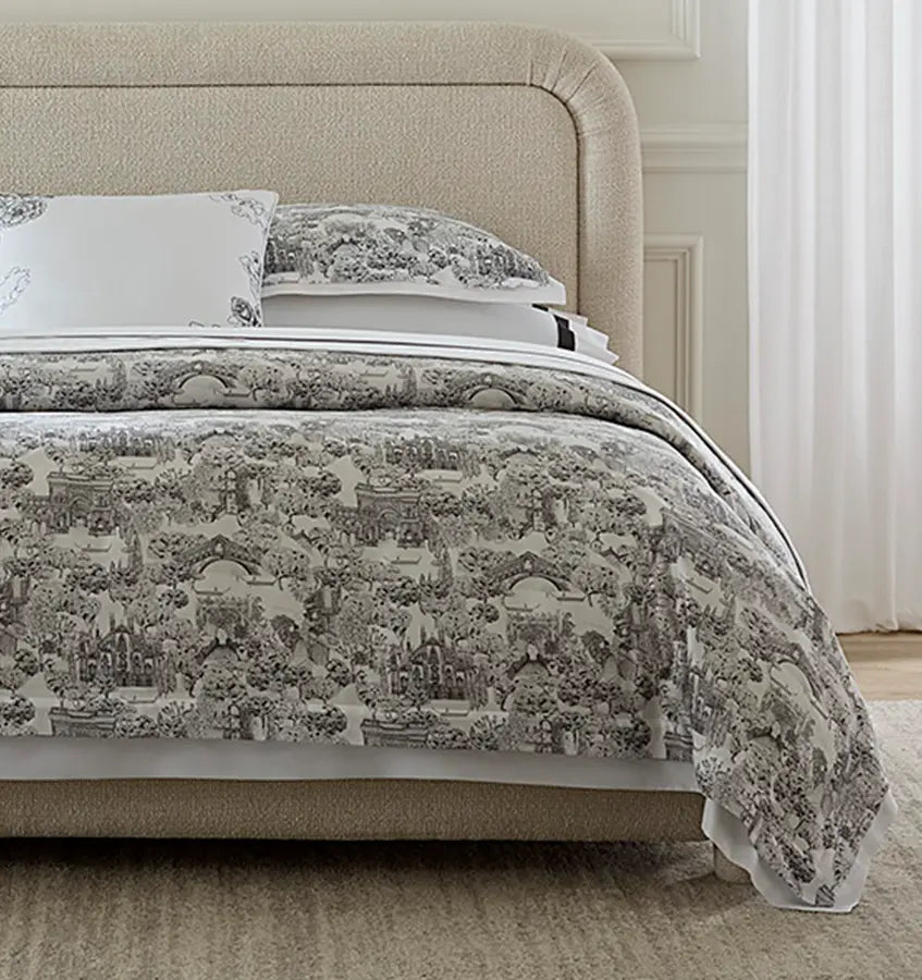 Charcoal toile cityscape print across a white duvet, draped over an upholstered bed.