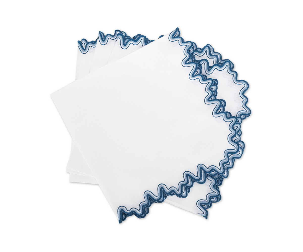 The Wave | Napkin by Matouk features three white linen napkins with blue scalloped edges, elegantly stacked and overlapping on a white background, adding a luxurious touch to your dining setting.