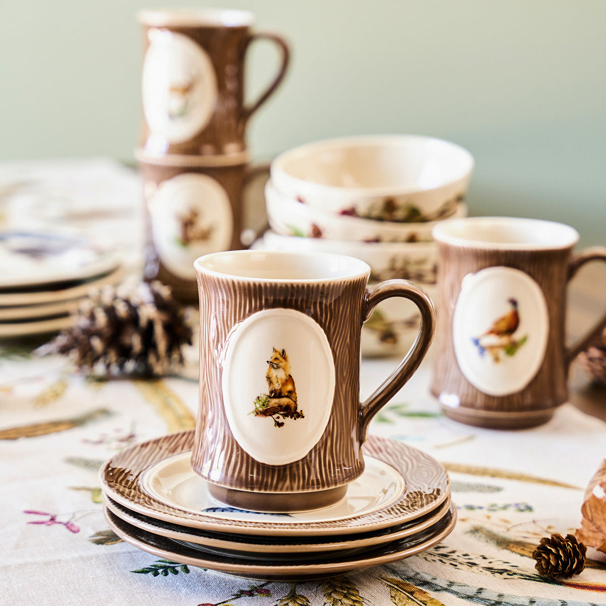 Forest Walk fox mug on stacked saucers with more animal mugs and pinecones on a patterned tablecloth