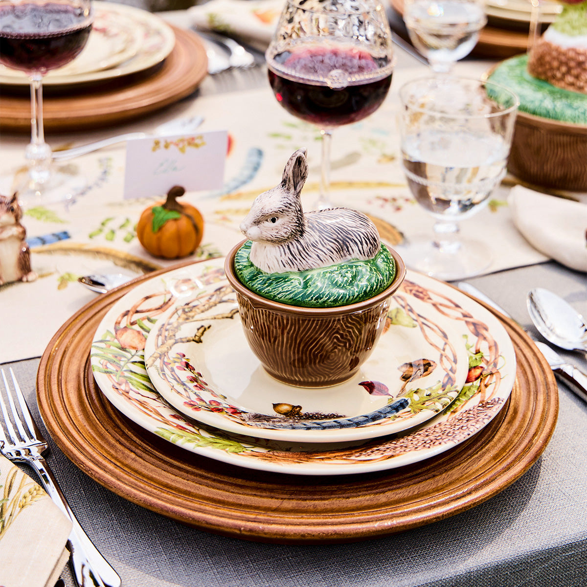 Forest Walk rabbit lidded bowl on stacked plates and a wood charger, with wine glasses and a small pumpkin