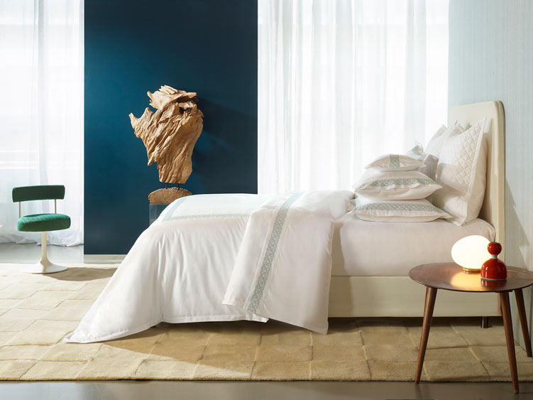 A stylish bedroom with the Elo Boudoir by Amalia Home Collection showcases a white bed with pillows, a round wooden side table with lamp and red vase, a green chair, and a wood wall sculpture on a dark blue accent wall.