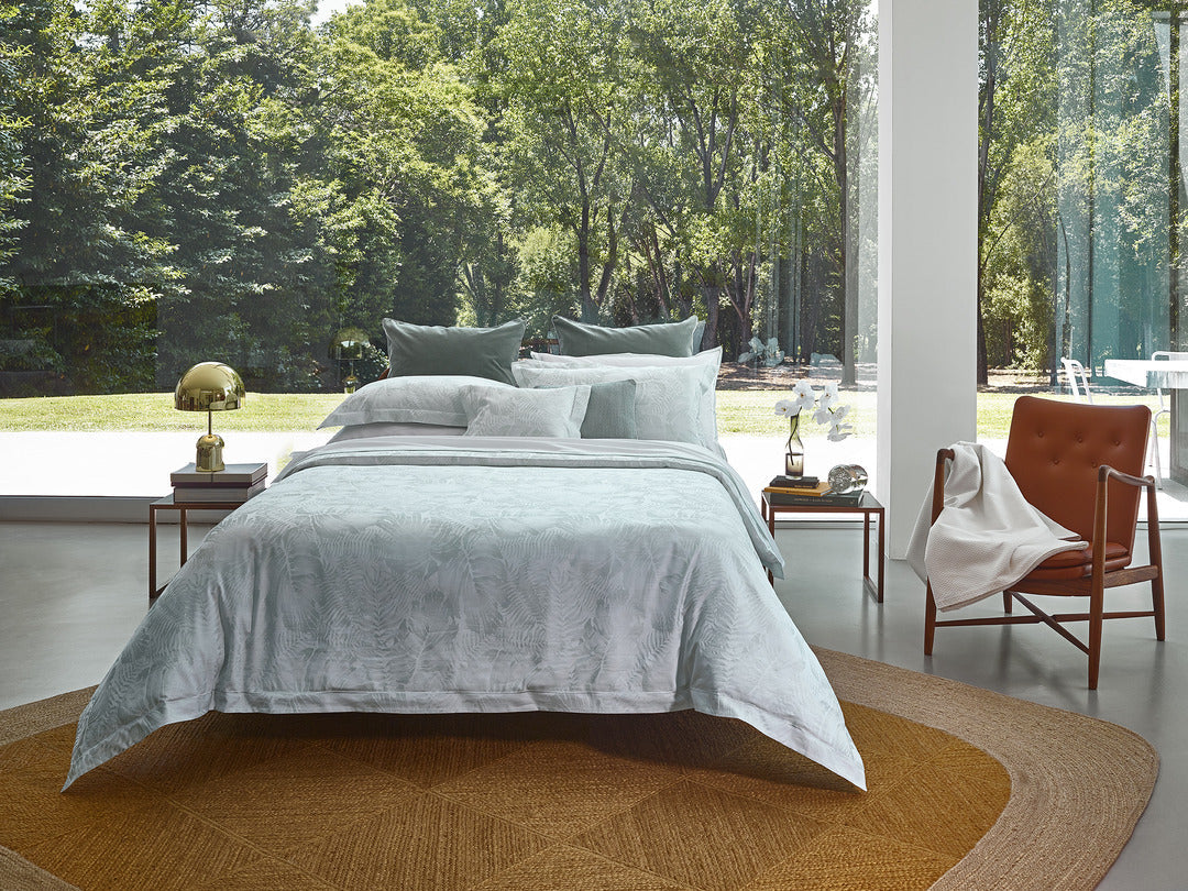 Sage green duvet cover styled on bed with accent pillows, floor-to-ceiling windows, and garden view.