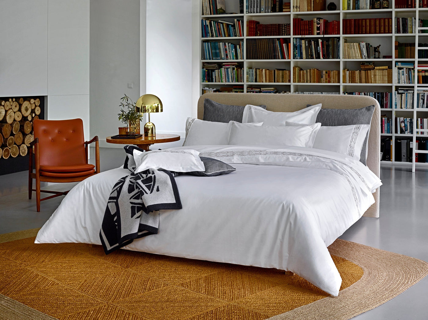 Styled bedroom with white flat sheet on made bed, library and leather chair in background.