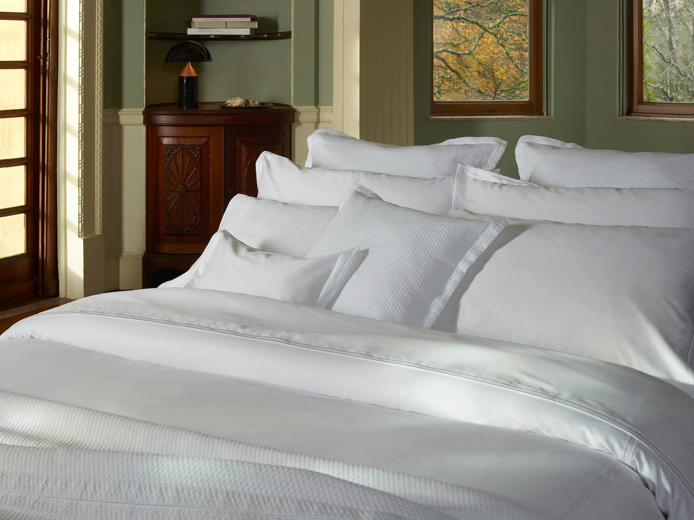 A sunlit bedroom with green walls and windows features a neatly made bed dressed in Sferra's Sereno Flat Sheet, crafted from crisp white Egyptian cotton sateen, complemented by multiple pillows.