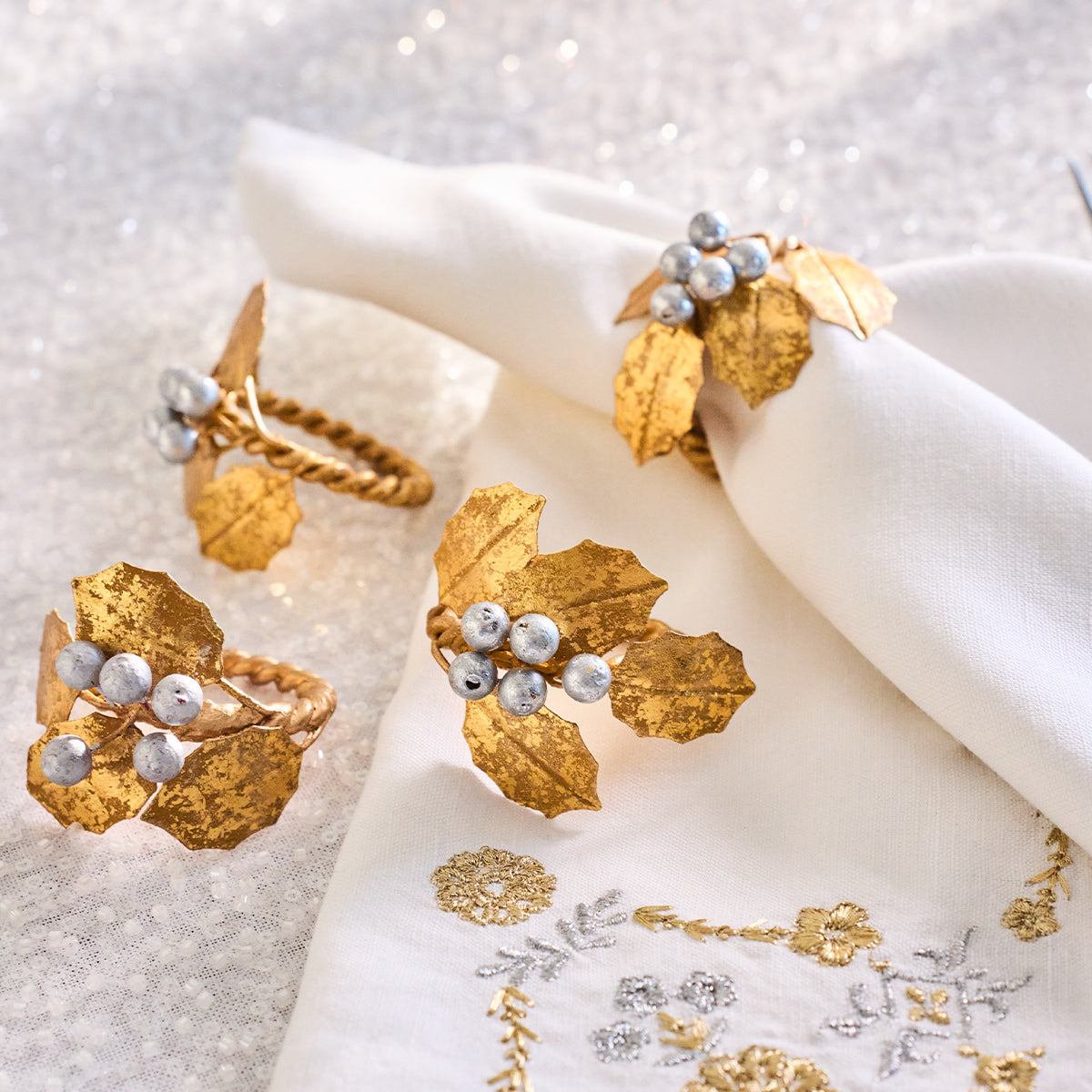 Gold holly napkin rings with silver berries around white napkins on a sparkling cloth